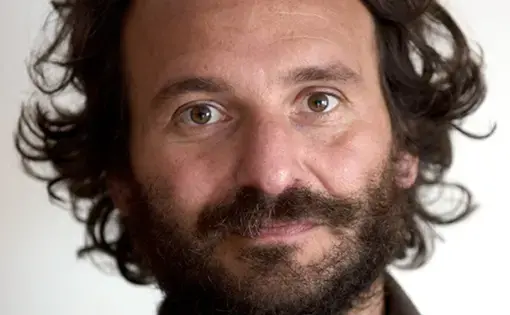 A man with curly brown hair and facial hair wears a brown button down