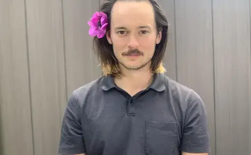 A man with longer brown hair wears a pink flower in his hair and a grey collared shirt