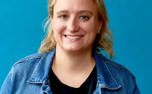 A blonde woman wears a jean jacket against a blue background