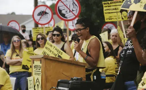 A woman wearing a yellow tank top speaks at a podium among a group of protestors, also wearing yellow