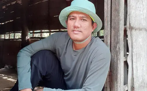 Suryadi sits on a threshold wearing a bucket hat.