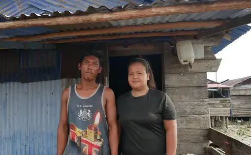 A man and woman stand together smiling against a decrepit house.