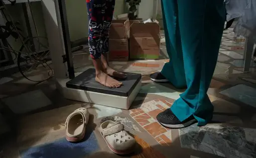Aimed at the feet of subjects, a child stands on a scale as a person wearing medical scrubs and tennis shoes faces them.