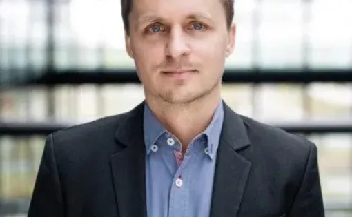 A man with short hair wearing a blue button down with a black blazer
