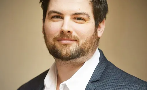 A man with short dark hair wearing a collared shirt and jacket against a beige background