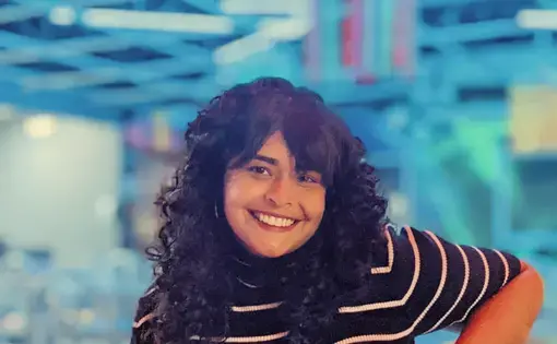A woman with dark curly hair wearing a striped shirt against a bright blue-lit background