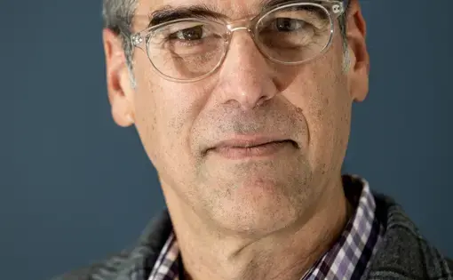 A man with short hair, glasses, wearing a plaid button down under a blazer