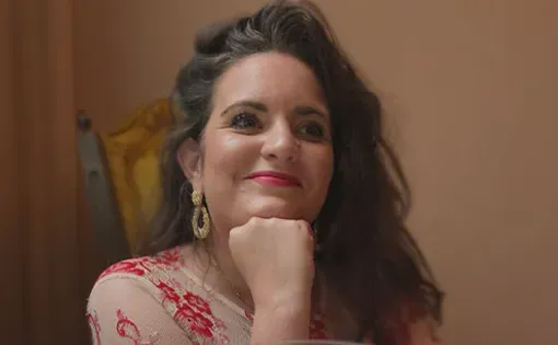 A woman with long dark wavy hair rests her chin on her fist while seated at a table