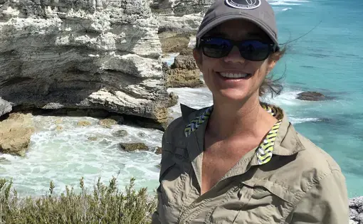 A woman wearing a baseball cap and green button-down against a background of cliffs beside the ocean