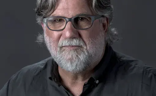 A man with grey hair, a beard, and glasses wearing a grey button-down