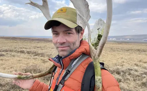 A man beside a large antler bone wearing an orange coat and green baseball cap