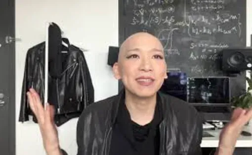 A person with a bald head wearing a leather jacket sits at a desk and gestures with both hands