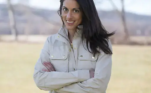 A woman with long dark hair stands in a field wearing a white coat with her arms crossed