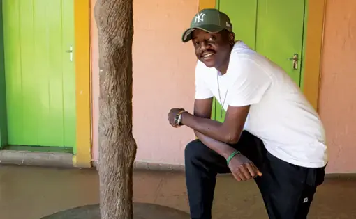 A man poses for a portrait. He wears a white t shirt and a baseball cap.