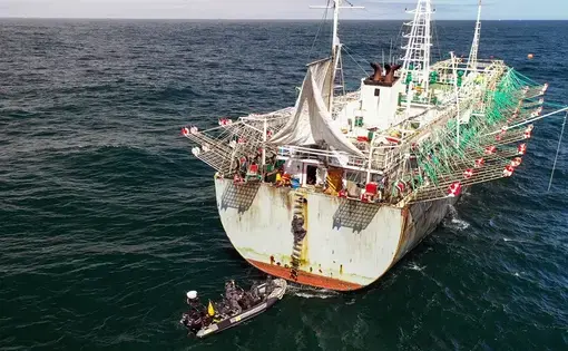 A squid jigger at sea during the day. Men in an inflatable boat have attached a rope ladder to the stern and have begun to climb aboard.