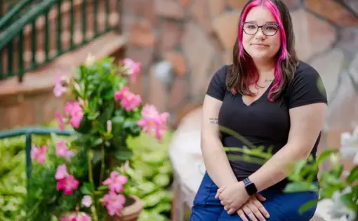 A young woman sits on a low wall in a garden blooming with pink roses