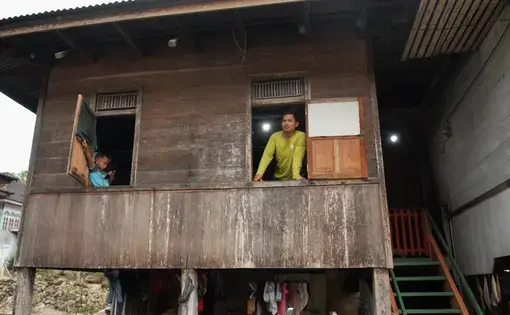 A man in yellow and a boy in blue stand in windows of a shack.