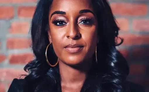 Headshot of a woman with long black hair wearing a black top and gold hoops, against a brick backdrop