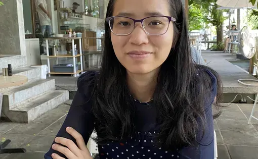A woman with dark hair and glasses sits at a table with her arms crossed