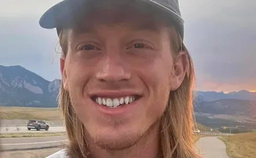 A man with shoulder-length blond hair wearing a dark blue baseball cap and grey sweatshirt against a grassy landscape