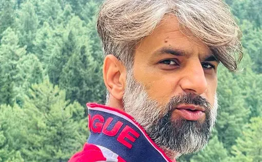 A man with grey hair and a beard wears a red and white striped shirt against a wooded background