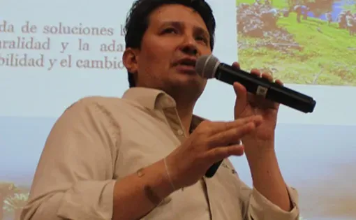 A man wearing a light button-down shirt holds and speaks into a microphone with slides projected behind him