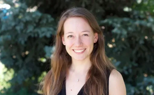 A woman with long brown hair wearing a black top standing against a tree