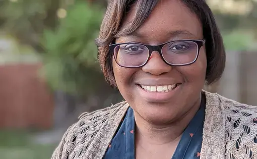 A smiling woman with short dark hair and glasses wearing a blue shirt and tan cardigan