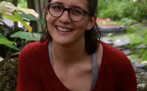 A woman with dark shoulder-length hair and glasses wearing a red t-shirt against a leafy background