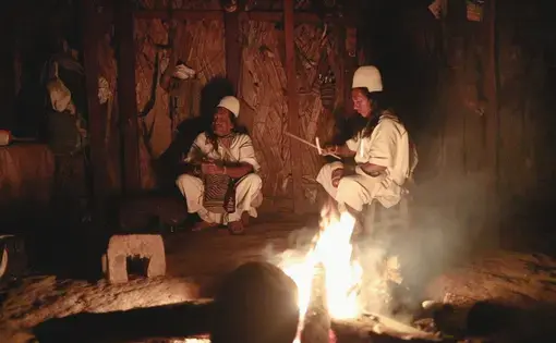 Two Arhuaco men dressed in white sit around a fire, which is in the foreground, at night