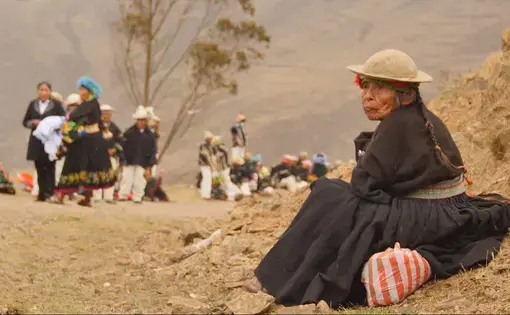 A Kallawaya woman wearing black with a tan hat sits on the ground in a mountainous area