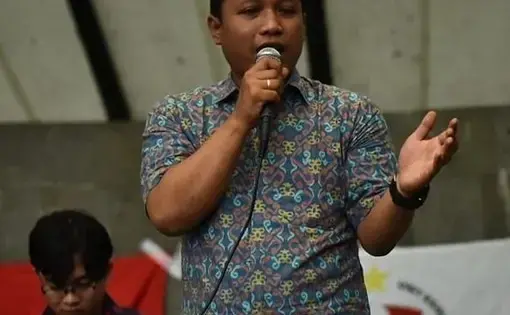A man in a short sleeve button down speaks into a microphone on a stage