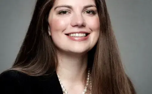 A woman with long dark hair wearing a black blazer against a grey background
