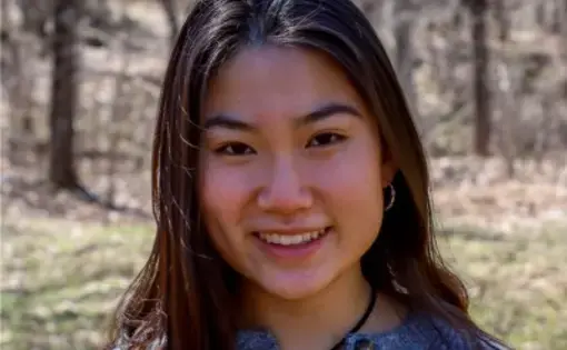 Headshot of a woman with long dark hair wearing a grey sweater against a wooded background