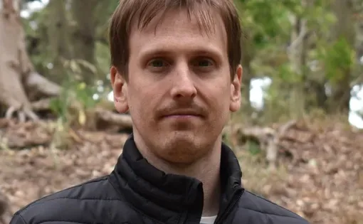 A man with short brown hair wearing a black puffer coat in a wooded area