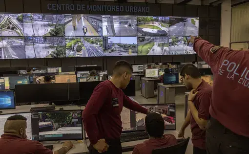 Police in t shirt uniforms sit in a room with monitors on the wall. Two men look at a computer screen and one points at a screen on the wall.