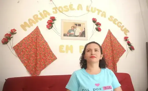 A woman in blue shirt sits and behind her is wall on which hangs a banner of José Cláudio and Maria's names.