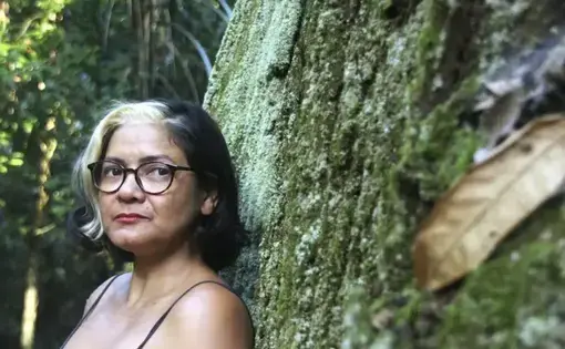 A woman with a streak of striking white hair leans against a giant tree and gazes into the camera.