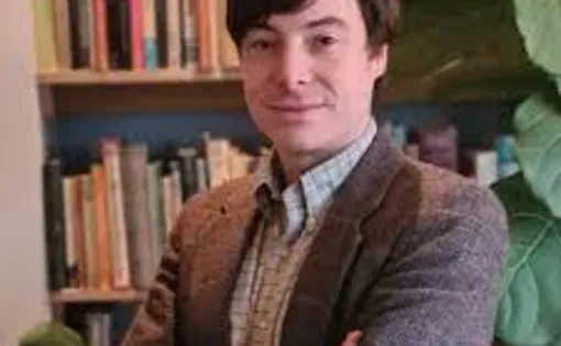 A man with short brown hair wearing a button down shirt and brown jacket stands against a bookshelf folding his arms