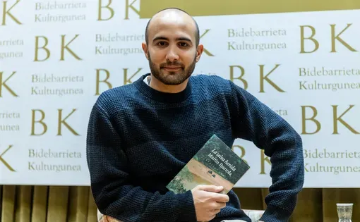 A man wearing a blue shirt sits holding a book in his arms.