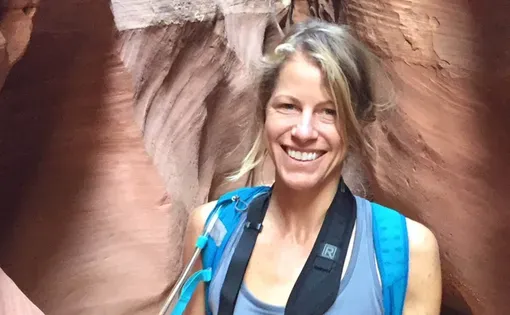 Carlyle wears a blue shirt and backpack while walking through a rocky formation