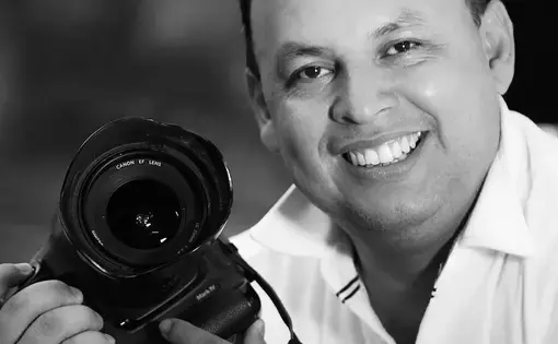 A man gleefully smiles at the camera while holding a camera in his hands.