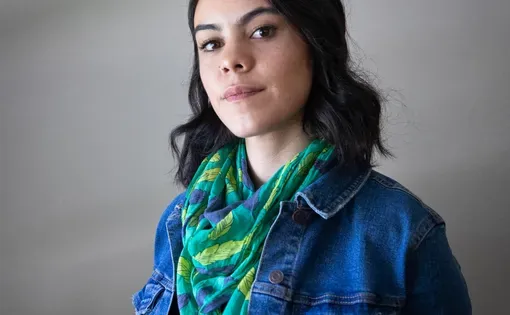 Headshot of Greta wearing a jean jacket and green scarf against a grey background