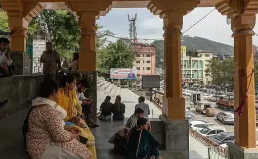 People sit on shaded stone steps lined by adorned columns, a cityscape in the background.