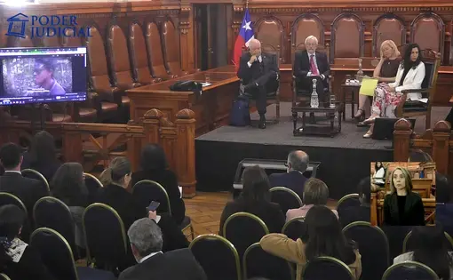 Four panelists sit on a stage wedged between rows of ornate seats lining the walls of a wooden-paneled room. They have an audience sitting in front of them and a television showing a video.
