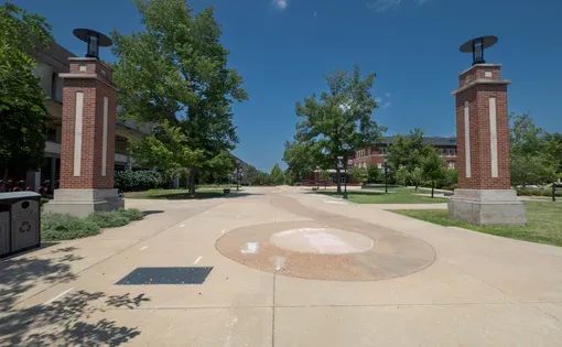 A stone plaza flanked by two brick columns. A green field and a building are in the background.
