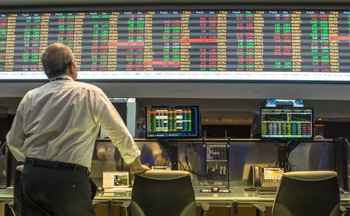 A man leans on his chair as he observes a large screen on the wall above with a string of red and green stock market values
