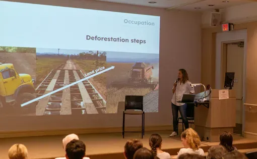 Fernanda stands in a lecture hall holding a microphone as she looks towards a projection of a presentation titled 'deforestation steps.' the pictures show a truck carrying lumber, a dirt road, and a soy combine, as a diagonal line goes up and right titled 'land price' indicating the value of the deforested land is increasing.
