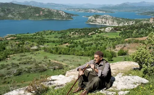 An elderly man sits atop a hillside overlooking a turquoise-blue river.