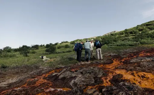 Scientists in the field gather near a spot on a grassy hill where rusty orange water and mud have accumulated.